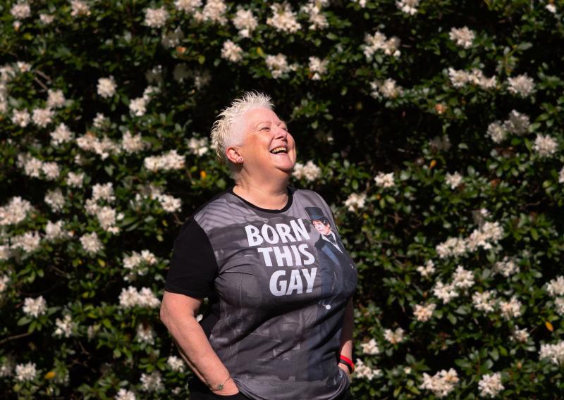 Older woman smiling wearing t-shirt "Born this Gay" against backdrop of flowers